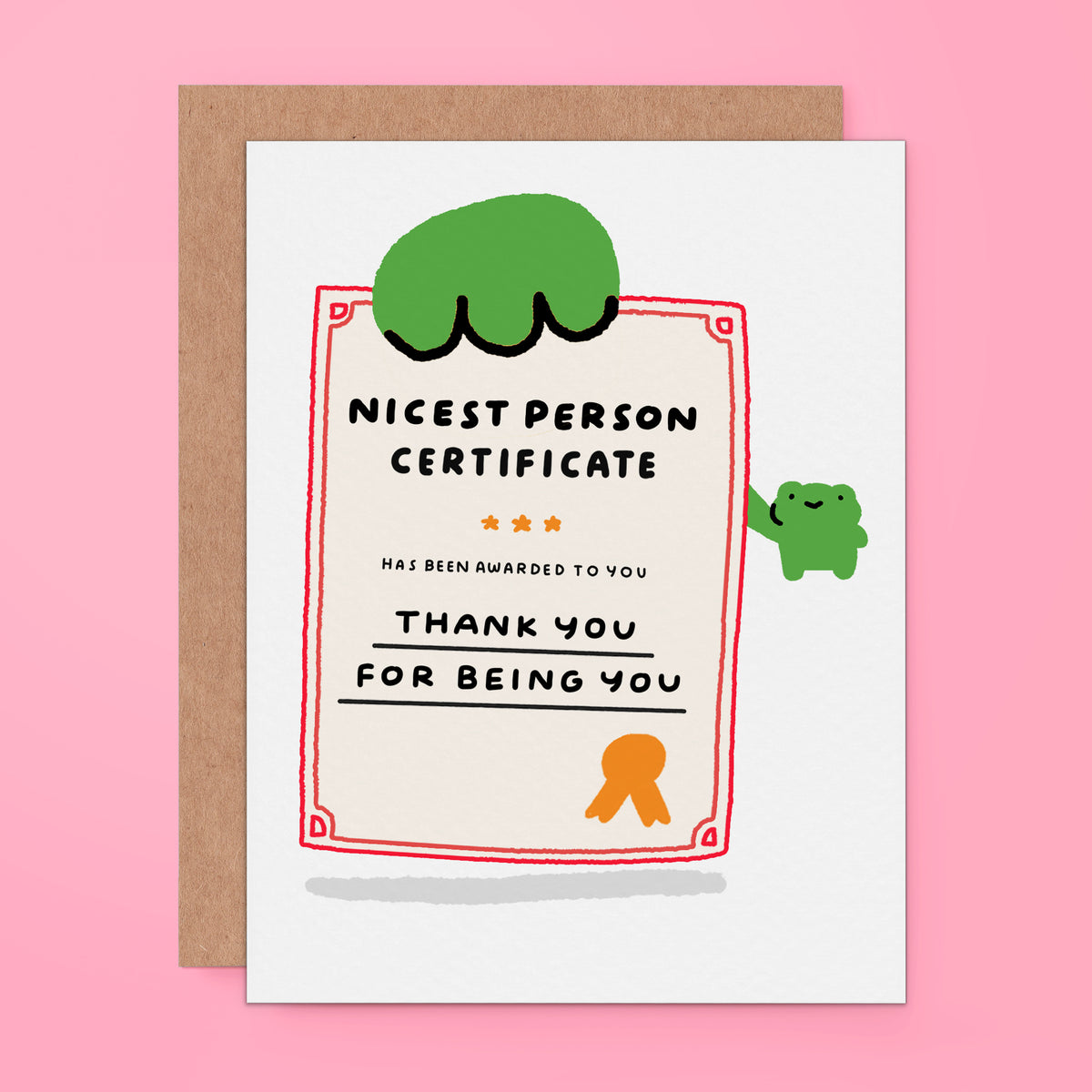 A novelty 'Nicest Person Certificate' card with a brown kraft envelope, displayed on a solid pink background. The card features a white base with a red border, black text, and cute cartoon frog illustrations in green and orange.