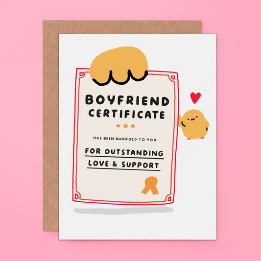 A novelty greeting card styled as a 'Boyfriend Certificate' with a brown kraft envelope behind it, set against a solid pink background. The card features a cartoon character and a small red heart.