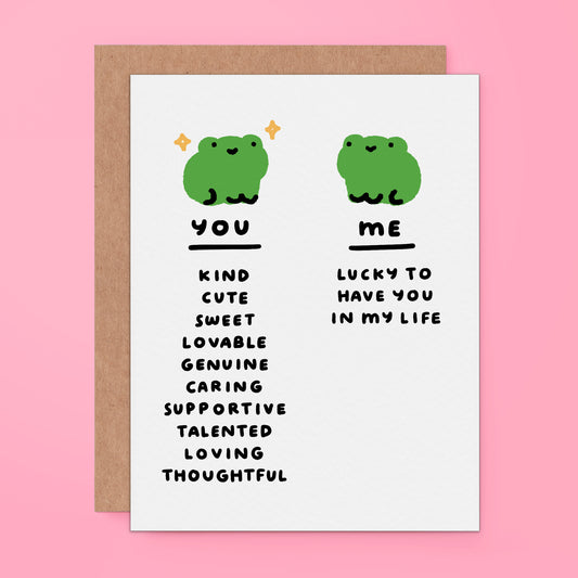A white greeting card with a brown kraft envelope behind it, featuring two green cartoon frogs and text comparing qualities of 'you' and 'me' on a pink background.